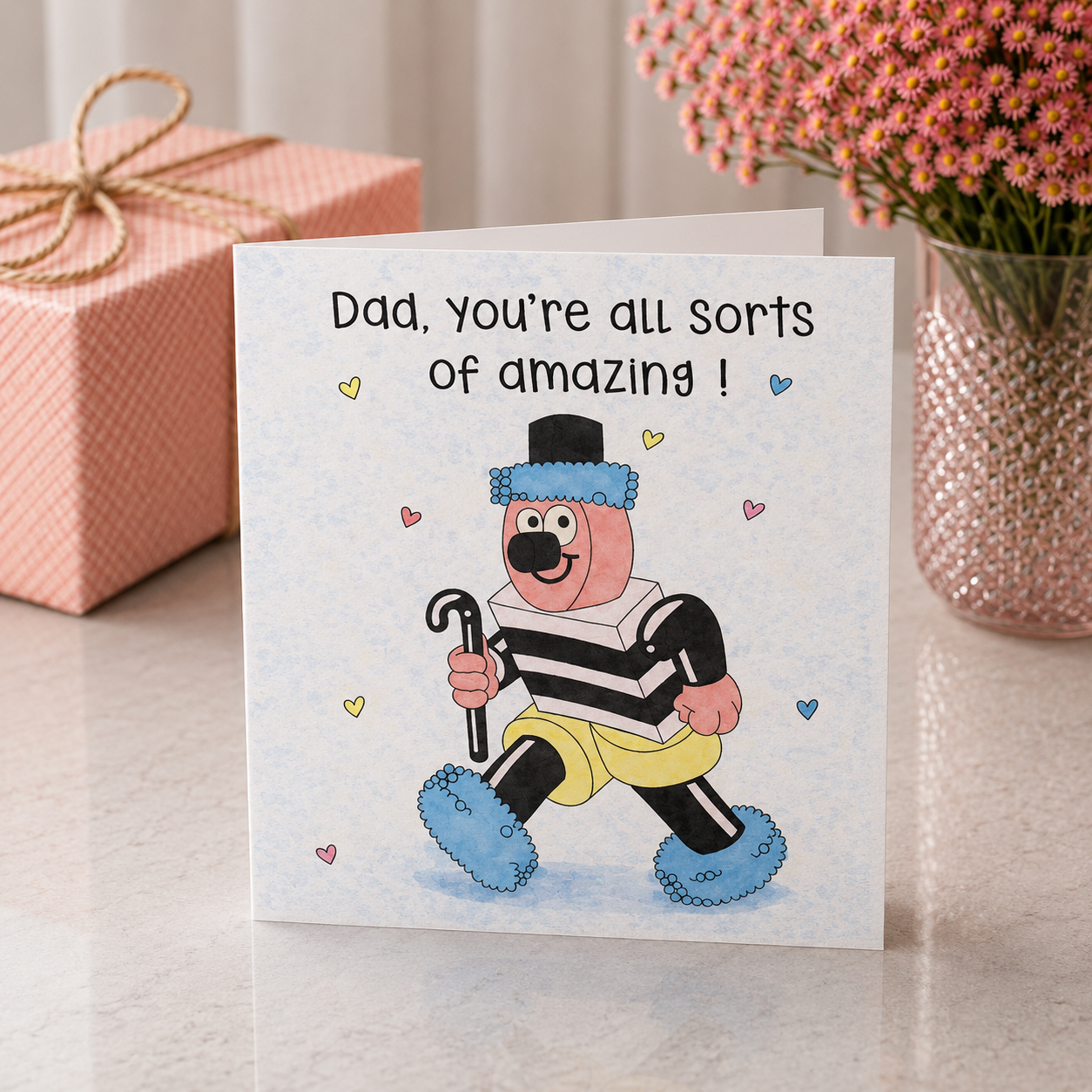 Greeting card with cartoon character and text 'Dad, you're all sorts of amazing!' on a surface with a gift and flowers in the background.
